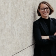 Freundlich lächelnde junge Frau mit Brille, halblangem dunkelblondem Haar und schwarzem Blazer lehnt an einer Wand, die Arme verschränkt.