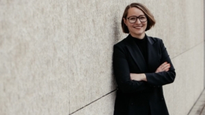Freundlich lächelnde junge Frau mit Brille, halblangem dunkelblondem Haar und schwarzem Blazer lehnt an einer Wand, die Arme verschränkt.