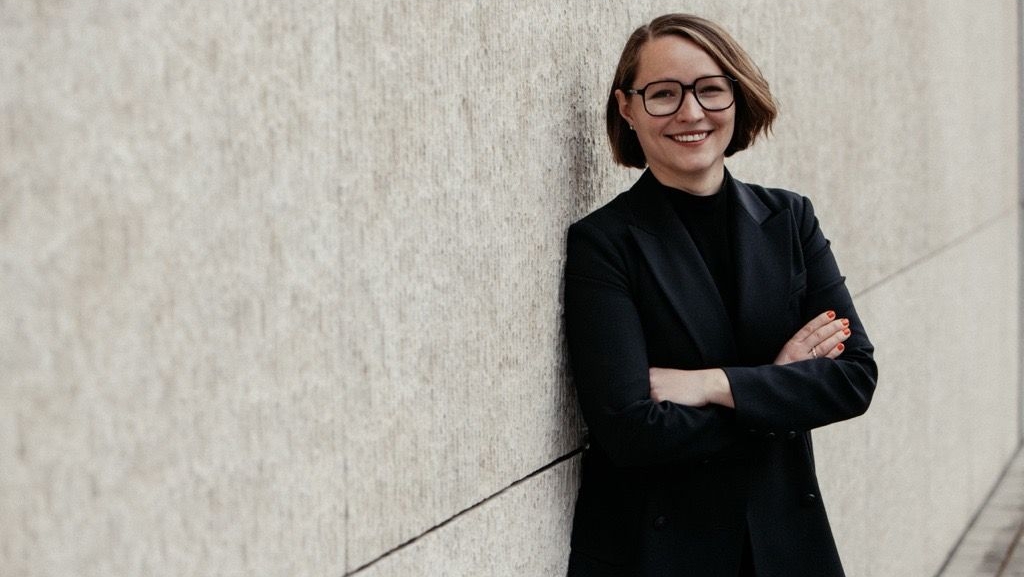 Freundlich lächelnde junge Frau mit Brille, halblangem dunkelblondem Haar und schwarzem Blazer lehnt an einer Wand, die Arme verschränkt.